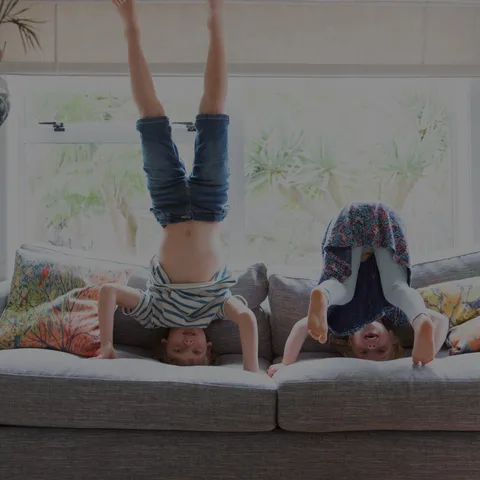Symbolbild: zwei Kinder machen spielerisch einen Kopfstand auf einem Sofa